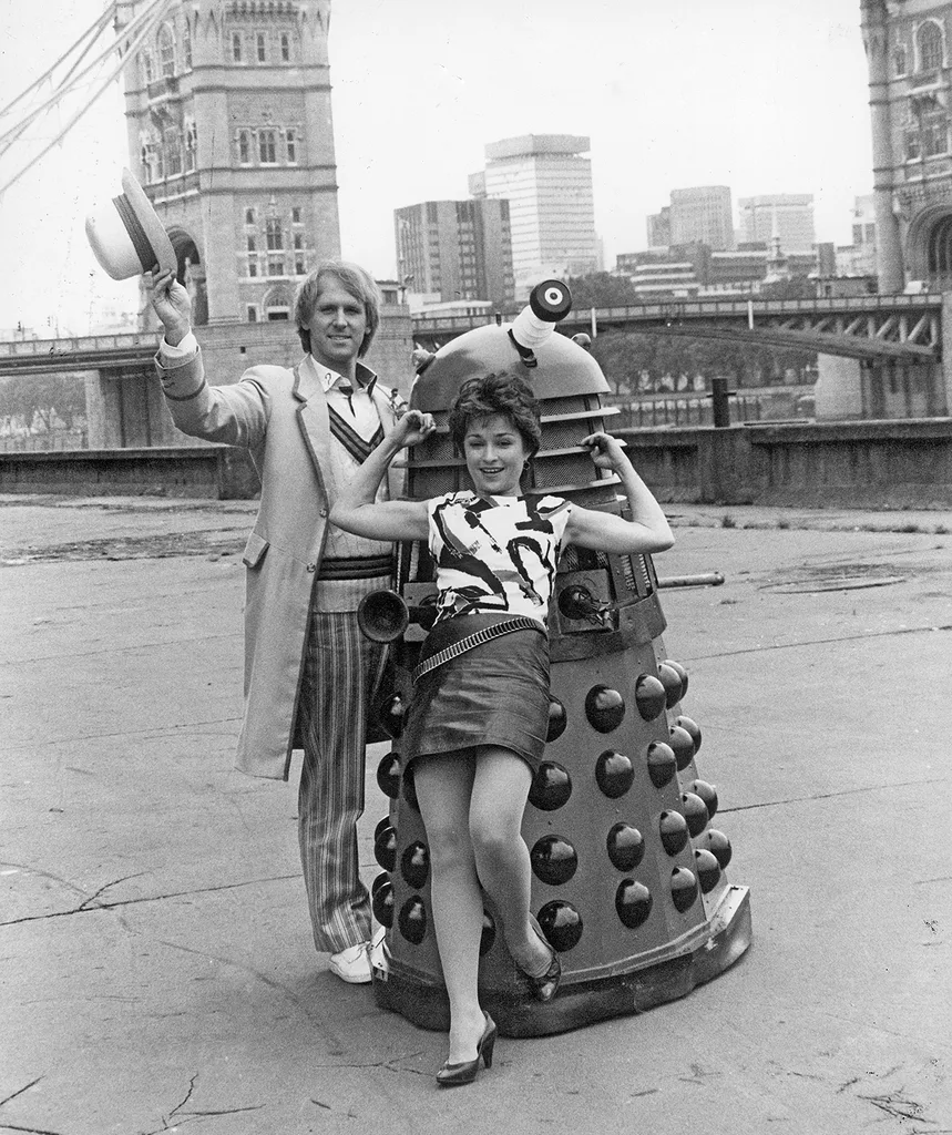 Actor Peter Davison who plays the "Fifth Doctor" in the BBC television science fiction series Doctor Who, seen here with actress Janet Fielding who plays his assistant in the series. They are seen in front of Tower Bridge with one of the Doctor Who Daleks. 13th September 1983. (Photo by Larry Ellis/Mirrorpix via Getty Images)