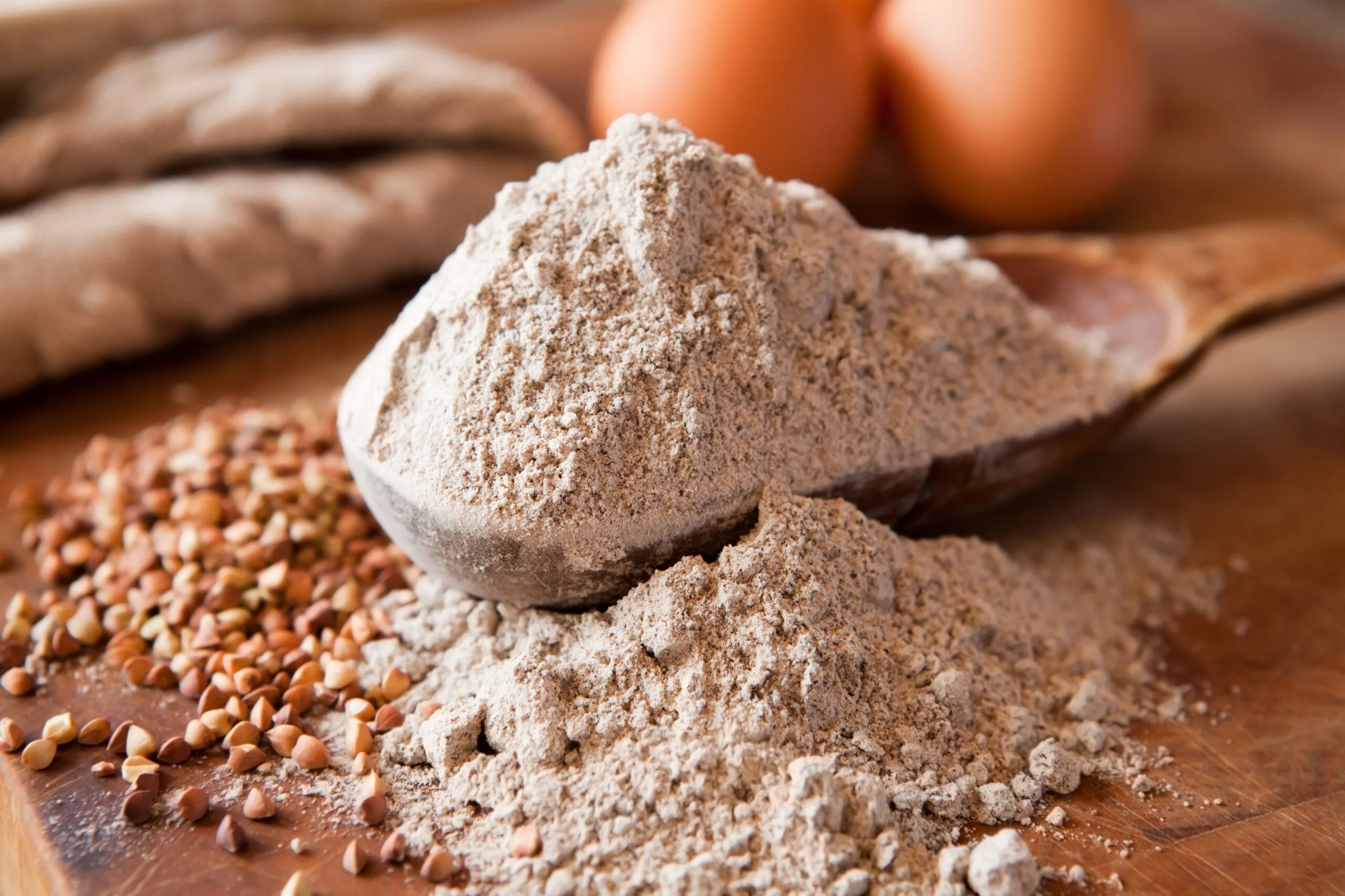 A wooden spoon filled with buckwheat flour with other grains and eggs nearby