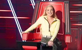 Tones and I, wearing a yellow top, sitting in one of The Voice's big red chairs