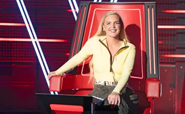 Tones and I, wearing a yellow top, sitting in one of The Voice's big red chairs