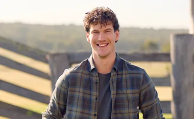 Zac, in a checked shirt, stands in front of a farm fence