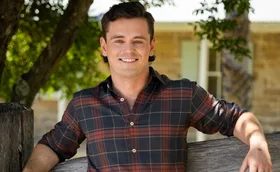 Alex, with a checked shirt on, smiles while standing outside