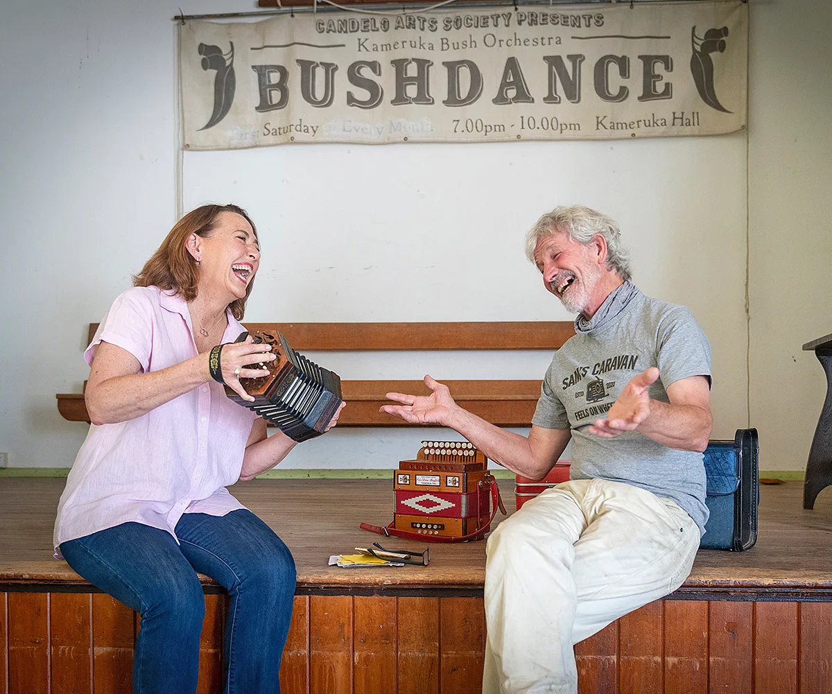 Lisa, holding an accordion, sits next to a man in front of a sign reading "bushdance"