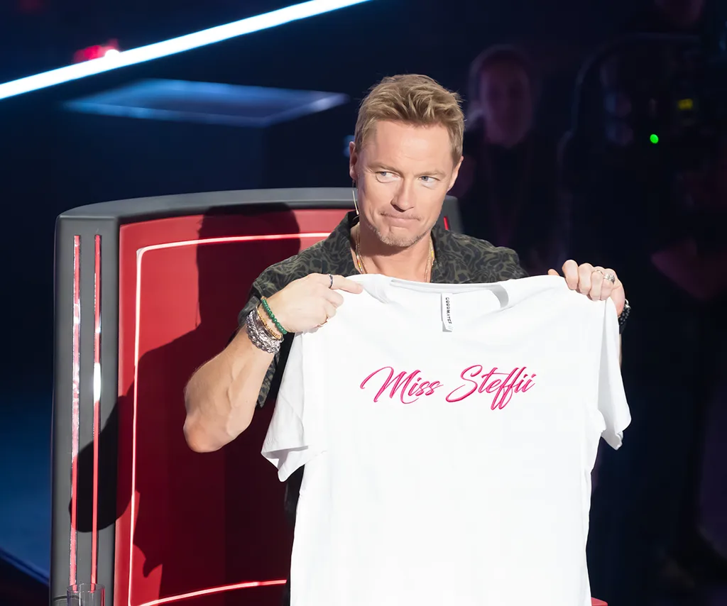 Ronan, sitting in the red chair, holds up a t-shirt saying "Miss Steffii".