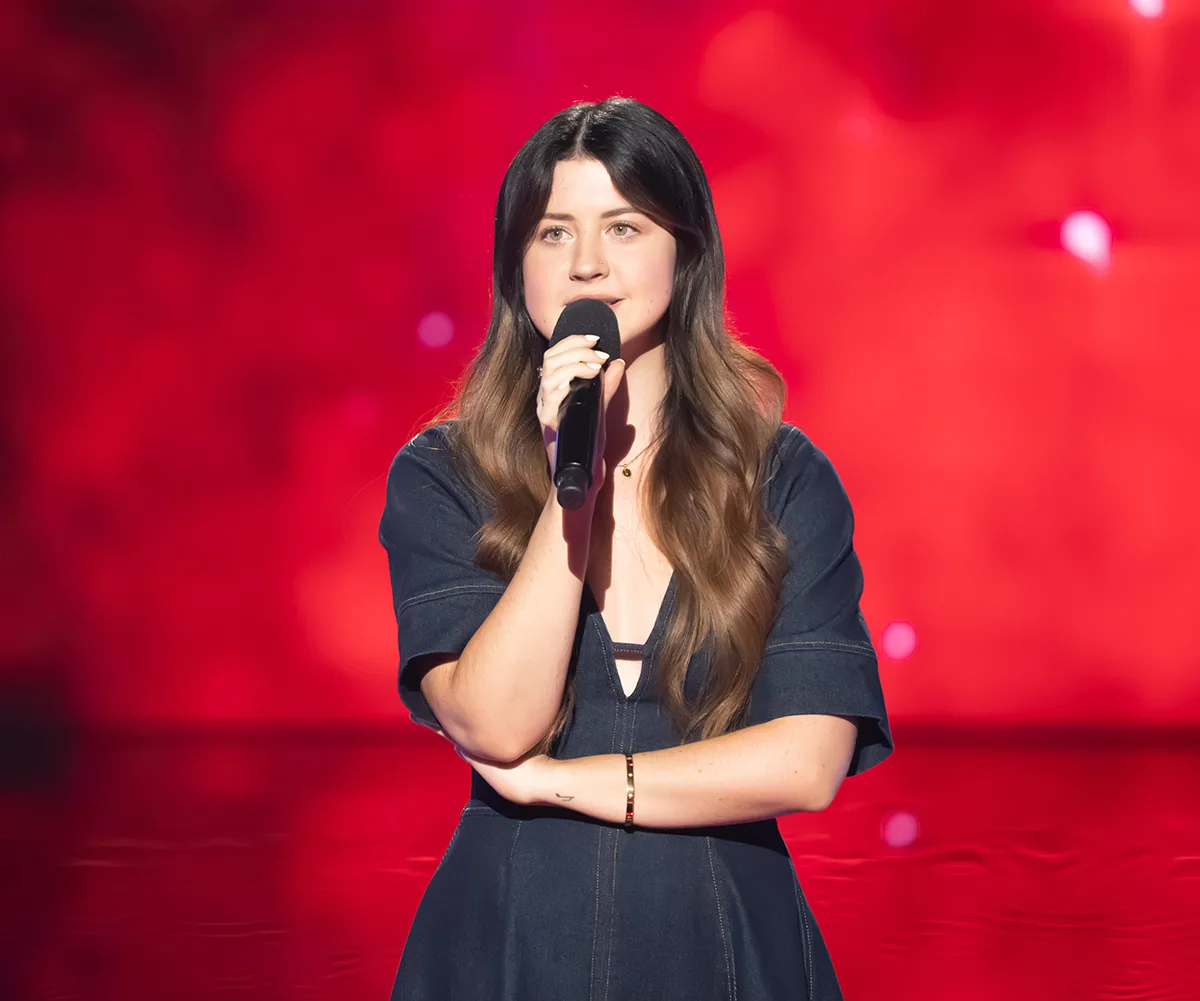 Charlotte stands on the set of The Voice in front of a red background.