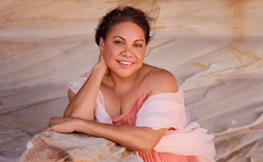 Deborah Mailman, in a pink dress, poses in front of a rocky background
