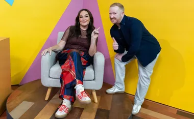Luke McGregor leans towards Kate Langbroek sitting in a chair on set