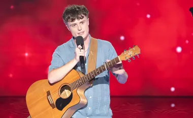 Findlay holds a guitar and sings in front of a red background
