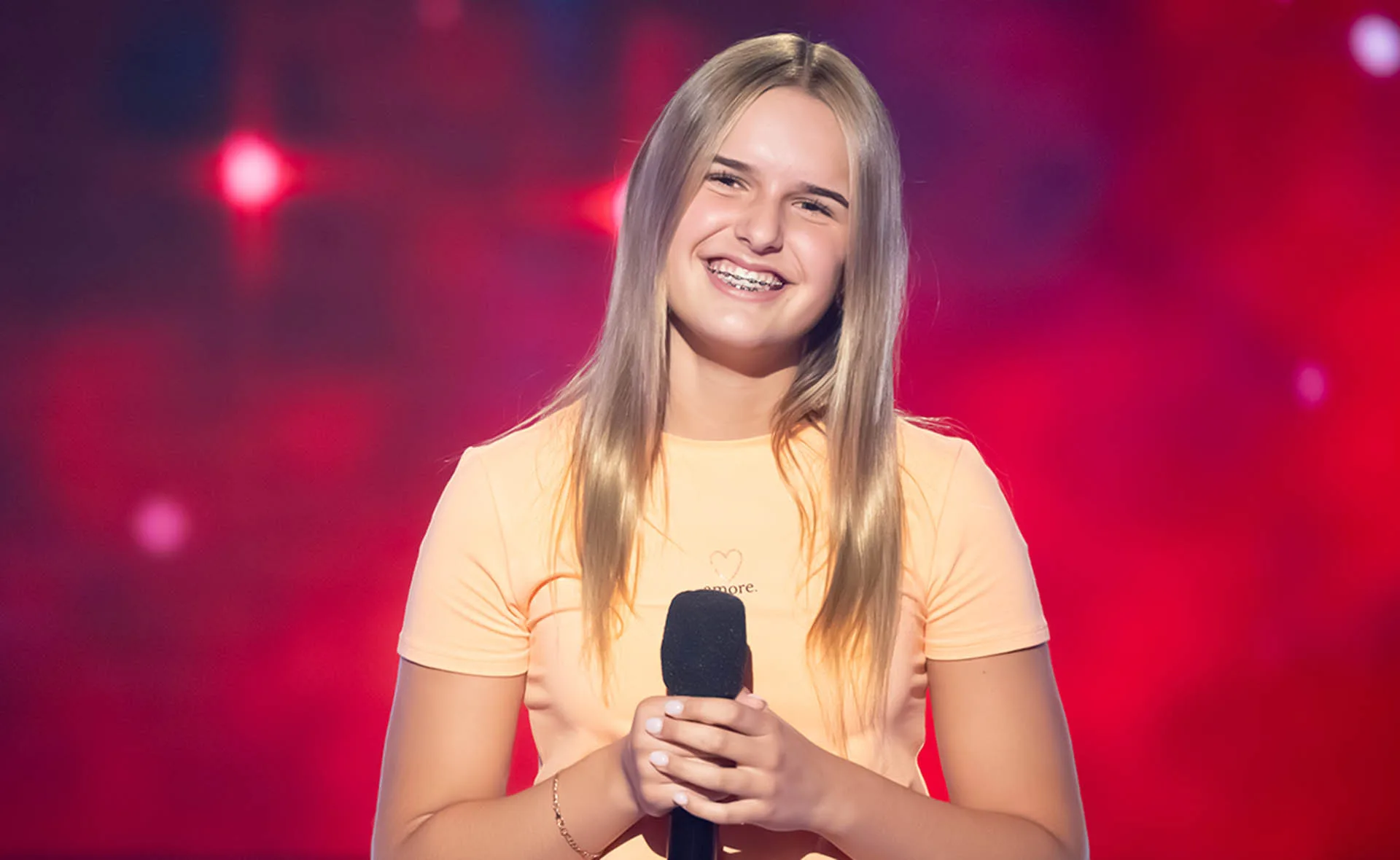 Leyla, wearing an orange t-shirts, smiles while holding a microphone on the set of The Voice