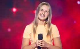 Leyla, wearing an orange t-shirts, smiles while holding a microphone on the set of The Voice