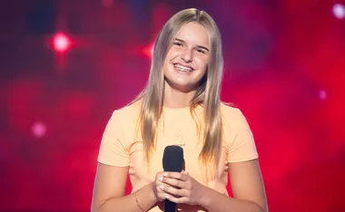 Leyla, wearing an orange t-shirts, smiles while holding a microphone on the set of The Voice