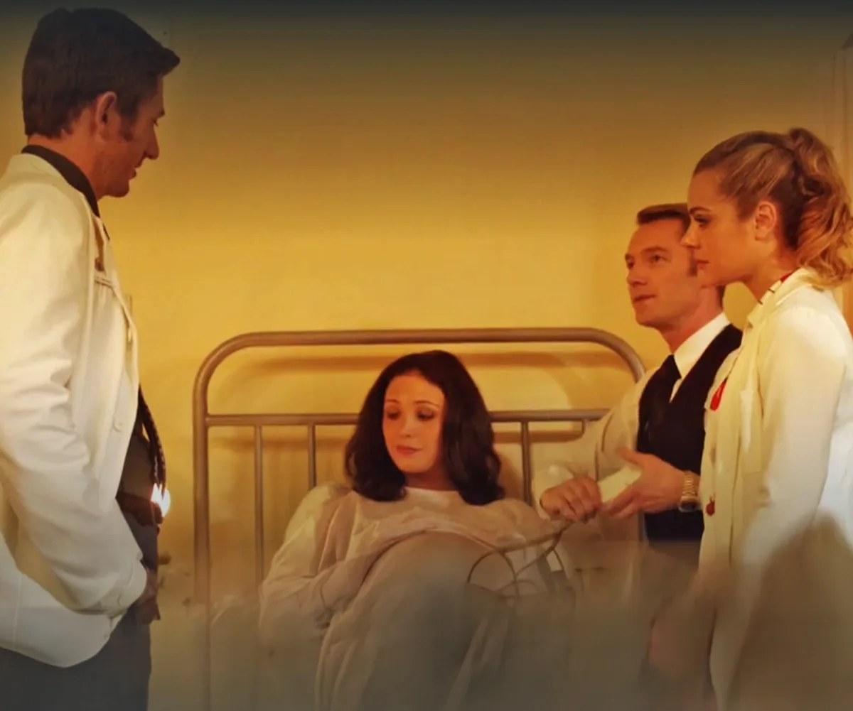 Taylor, in character, lies in a hospital bed, with Ronan, playing a doctor, standing next to her, along with two other doctors