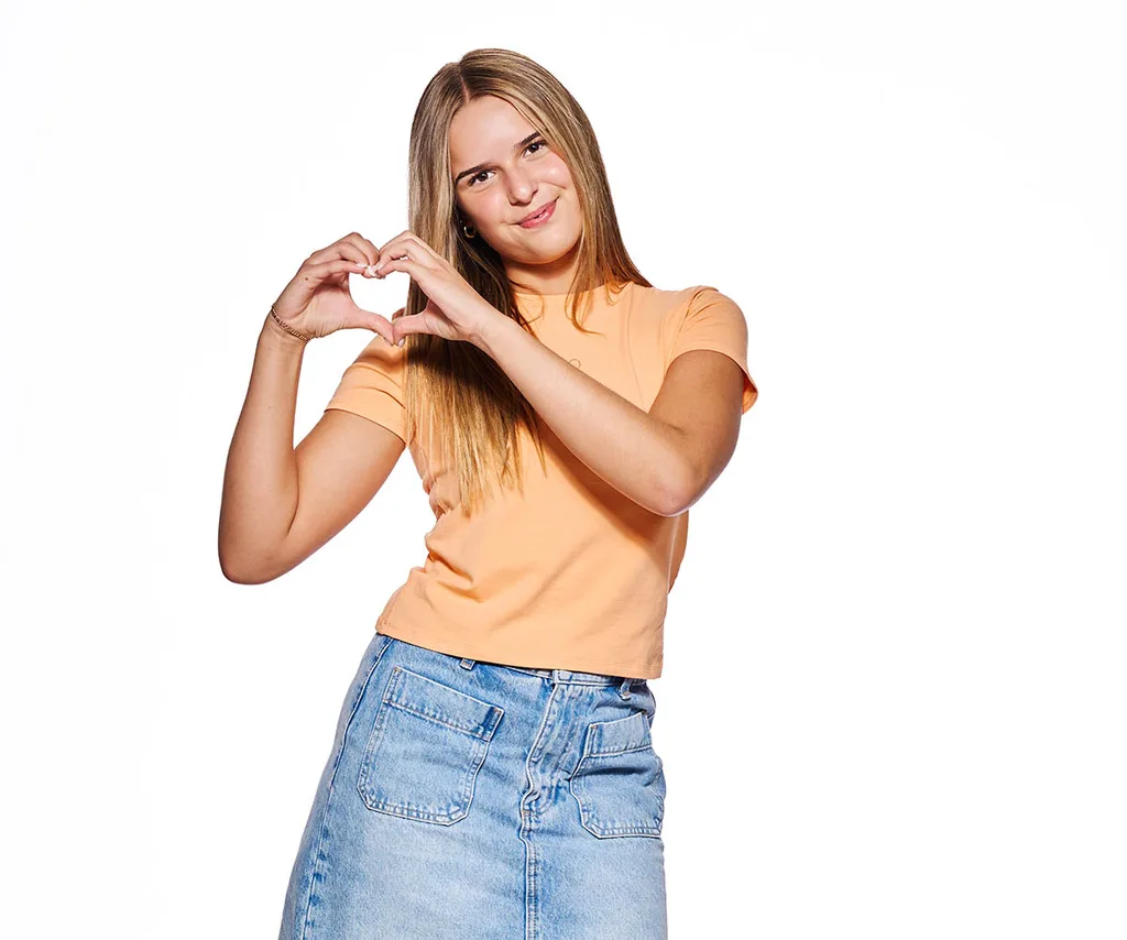 Leyla, wearing an orange t-shirt, makes a heart shape with her hands
