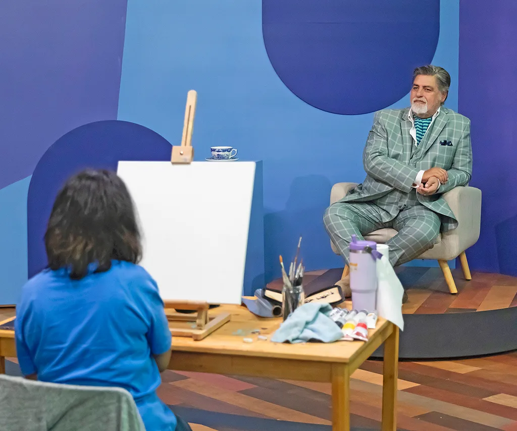 Matt sits on the set of Portrait Artist of the Year, with an artist painting his portrait in the foreground