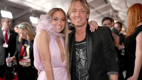 JLo in a pink dress and Keith Urban in a tshirt and black jacket, with JLo's arm around Keith's shoulders. Both are smiling on the red carpet.
