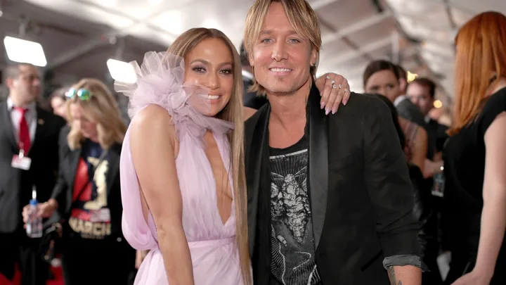 JLo in a pink dress and Keith Urban in a tshirt and black jacket, with JLo's arm around Keith's shoulders. Both are smiling on the red carpet.