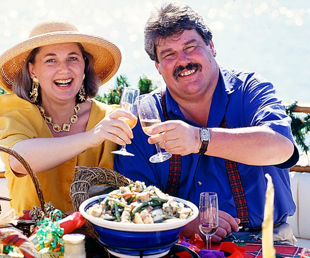 Tonia Todman, wearin a yellow dress and hat and Huey, wearing a blue shirt and red suspenders, both smiling and clinking champagne glasses on Healthy, Wealthy and Wise in the 1990s.