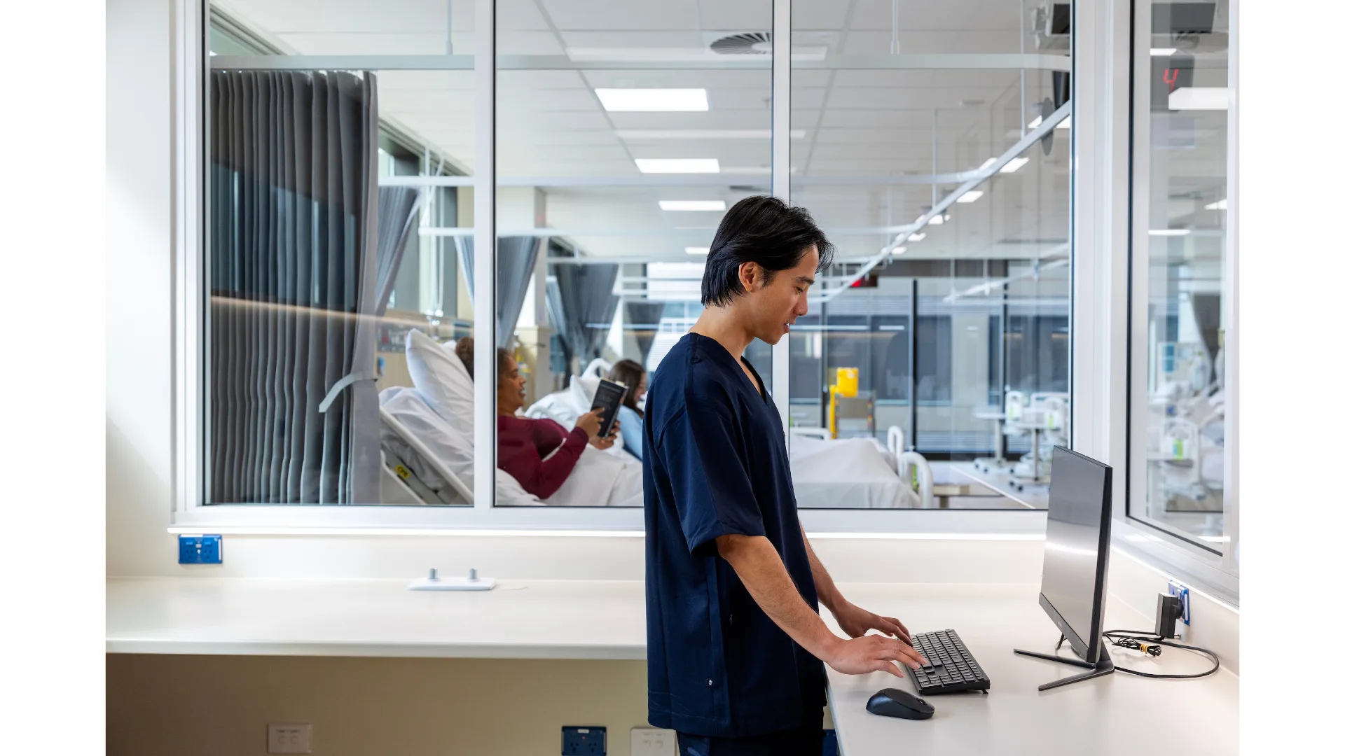 A medical professional works at a computer as female patients rest in beds in a ward.