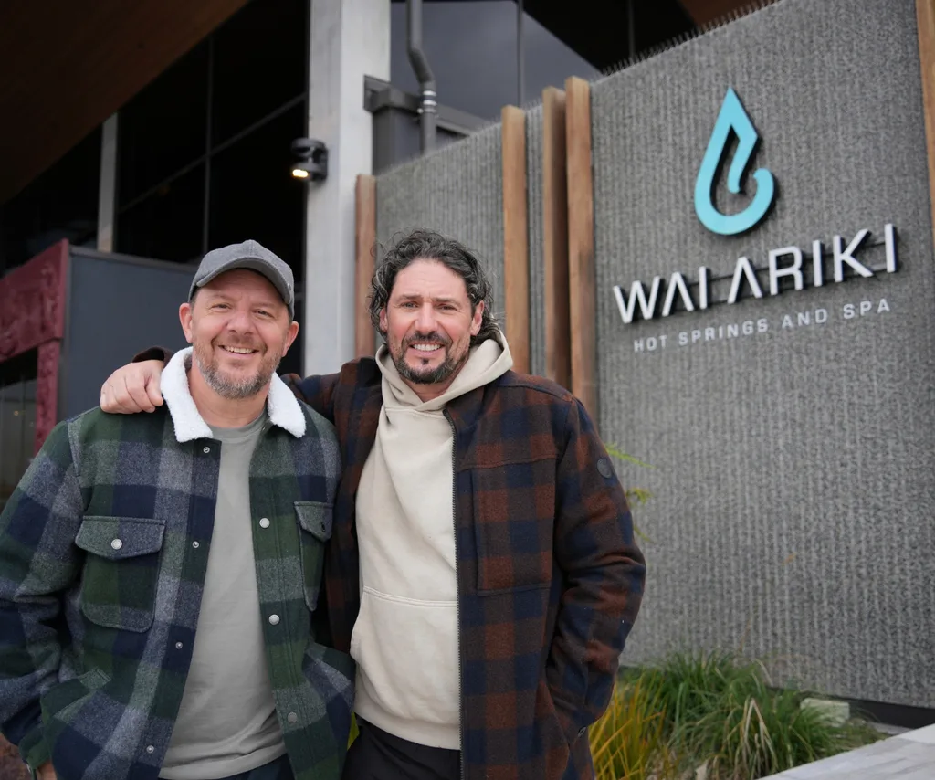 Colin stands with his arm around Manu in front of the entrance to a building