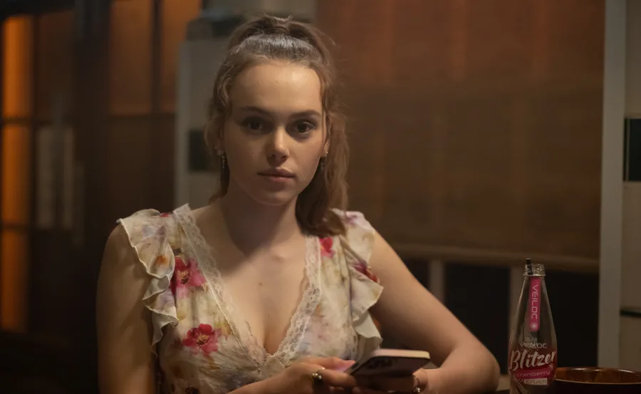 Zoe, wearing a red dress, sits in a bar with a drink in front of her, in her role as Edie