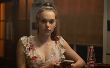 Zoe, wearing a red dress, sits in a bar with a drink in front of her, in her role as Edie