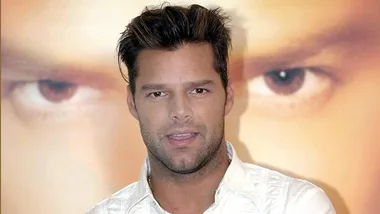 Ricky Martin in a white shirt stands in front of a large photo showing a close-up of eyes.