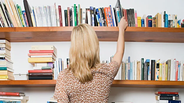 A woman reaches for a book on a wooden bookshelf filled with various books.