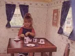 Child playing with a tea set at a small wooden table inside a cozy room with blue curtains and floral wallpaper.