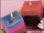 Square multi-colored candles with flames, set on a pink surface, alongside a few petals.