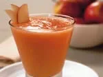 A glass of orange smoothie garnished with apple slices, with a bowl of apples blurred in the background.