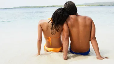 A couple sits on a sandy beach, backs to the viewer, with a serene sea and distant hills in the background.