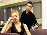 Man and woman appear tense in a modern living room, woman sitting while man stands holding a wine glass.