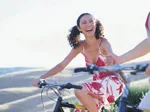 A woman joyfully rides a bicycle outdoors on a sunny day, wearing a floral dress, with rolling hills in the background.