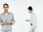 Woman in a suit holding a notepad, with a blurred person walking in the background against a plain white wall.