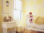 Yellow nursery with a crib, a doll wearing red, wall art, and a white dresser with a skirted stool.