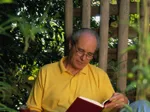 An older man with glasses is sitting outdoors, wearing a yellow shirt, and reading a red book in a garden setting.