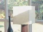 A lamp with a cube-shaped shade next to a window and a vase in the background.