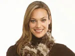 A woman wearing a brown turtleneck and a fluffy, multicolored knitted scarf smiles at the camera.