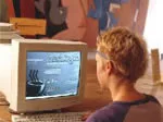 A child with short hair sits at a desk, playing a computer game on an old CRT monitor in a room with colorful walls.