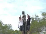 Two people stand on a statue pedestal outdoors, gesturing with arms outstretched, with trees in the background.
