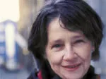 A woman with short dark hair, smiling, outdoors with a blurred urban background.