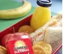 Lunchbox with a sandwich, orange juice, apple, crackers, and a banana in the background.