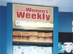 "Store display with 'Women's Weekly' magazine sign above shelves filled with magazines."