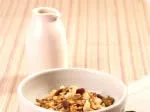 A bowl of granola with nuts next to a white ceramic milk jug on a light-colored, striped tablecloth.