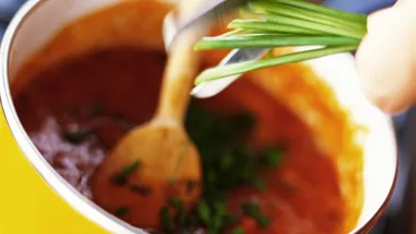 A pot of rich, red-orange stew being stirred, garnished with chopped herbs and sliced chives.