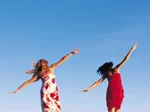 Two women in dresses joyfully leaping against a clear blue sky.