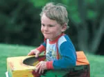 A young child outdoors with a painted box, wearing a colorful shirt, smiles slightly with greenery in the background.