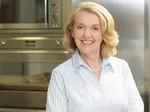 A smiling woman in a kitchen setting, wearing a light blue blouse, standing in front of modern stainless steel appliances.