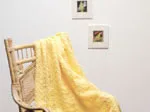 A yellow crochet throw rug draped over a bamboo chair, with two framed pictures on the white wall behind.