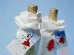 Two gift-wrapped bottles with festive tags featuring holiday-themed illustrations, one with a red dress girl and another with a red tree.